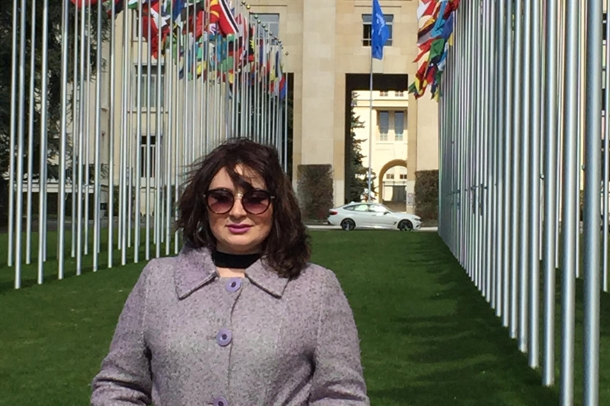 [ai] A woman wearing sunglasses and a purple coat stands on a grassy area surrounded by flagpoles displaying various international flags, with a building and a car visible in the background.